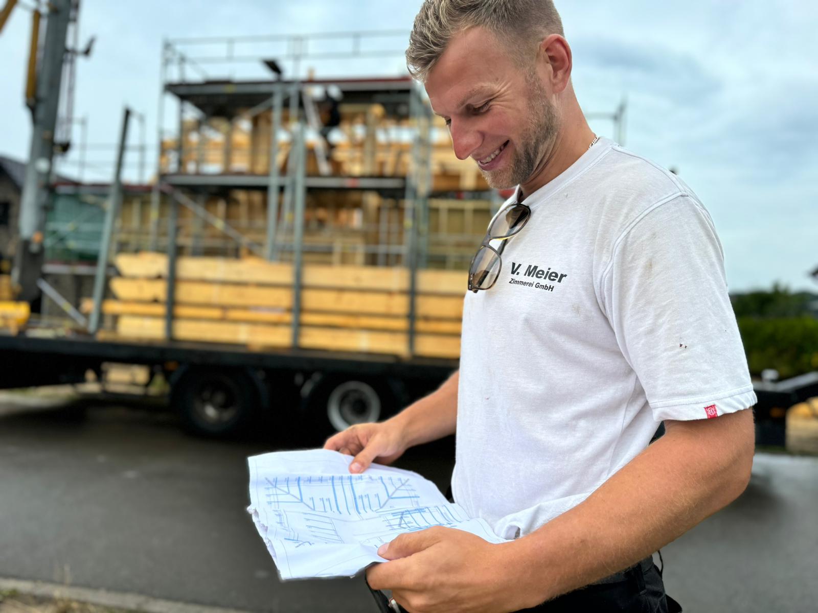 Lachender Zimmerer von V. Meier liest einen Bauplan vor der Baustelle